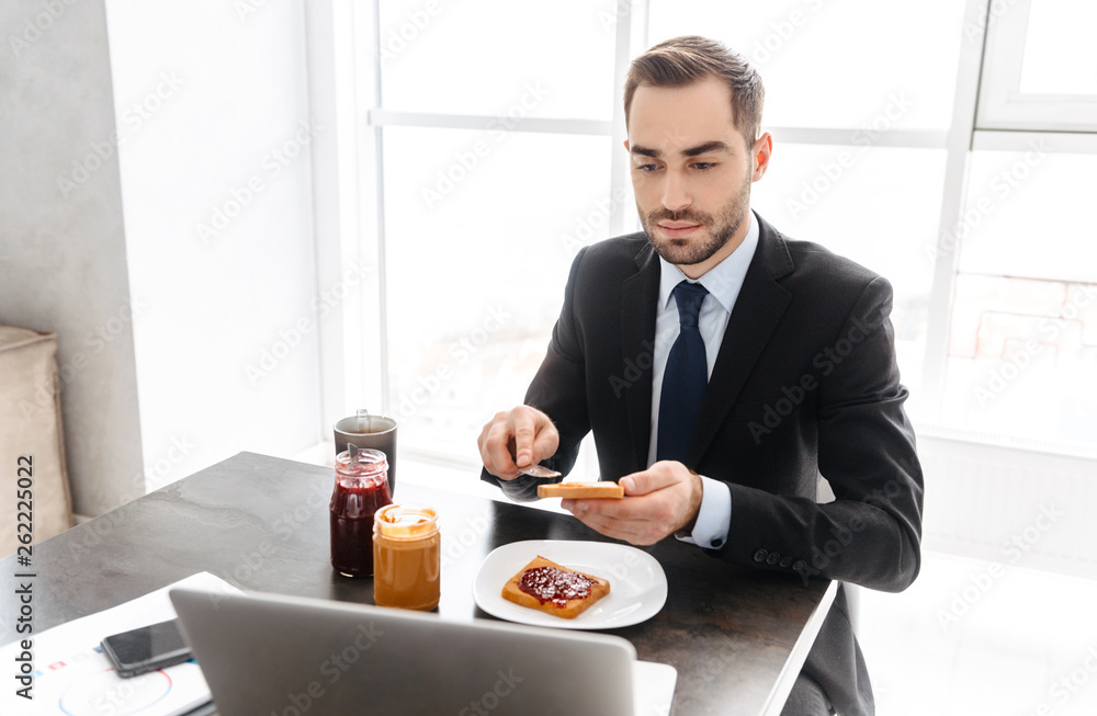 Image of businesslike man having breakfast while working on laptop in bright office Image of businesslike man having breakfast while working on laptop in bright office