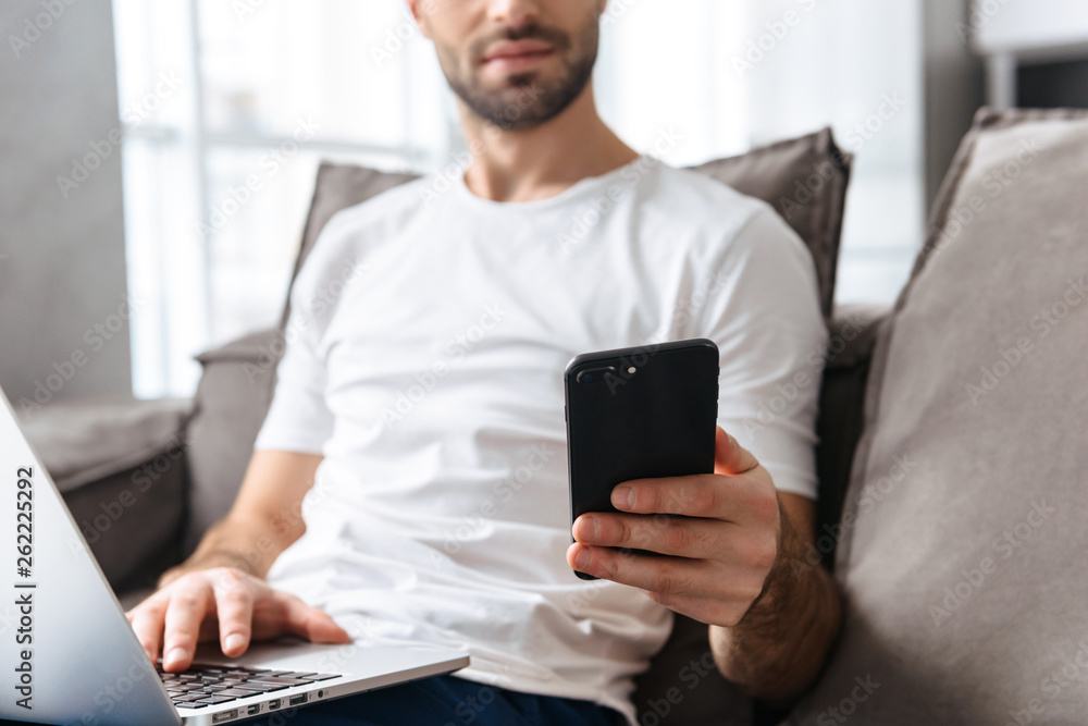 Photo of serious guy holding smartphone and using silver laptop while sitting on sofa in bright apartment