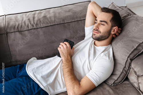 Photo of caucasian guy holding and using smartphone while lying on sofa in bright apartment