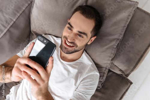 Photo of attractive guy holding and using smartphone while lying on sofa in bright apartment