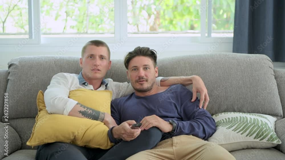 Young gay male couple relaxing on couch in living room watching tv. Caucasian, european men sitting on sofa. Scare mood. Happy gay friendship, relationship, lifestyle concept. Young gay male couple relaxing on couch in living room watching tv. Caucasian, european men sitting on sofa. Scare mood. Happy gay friendship, relationship, lifestyle concept.