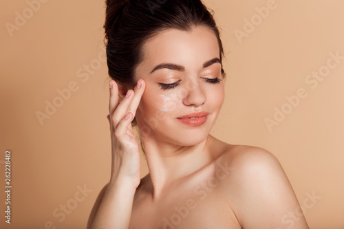Beautiful face portrait of young woman is applying face cream on a cheek. Skin care and health concept.