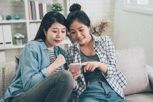 Front view portrait of two joyful asian female friends listening music on line from a smart phone sitting on couch in living room at home. young girls sharing earphones pointing show cellphone screen