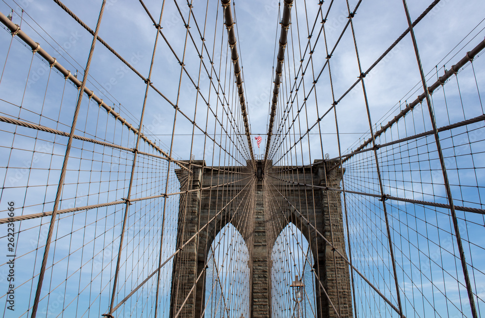 Fototapeta premium Słynny most Brooklyn Bridge w Nowym Jorku w Stanach Zjednoczonych Ameryki, przedstawiający zawieszenie przewodowe i flagę USA na szczycie kolumny w częściowo pochmurny dzień z niebieskim Skys.