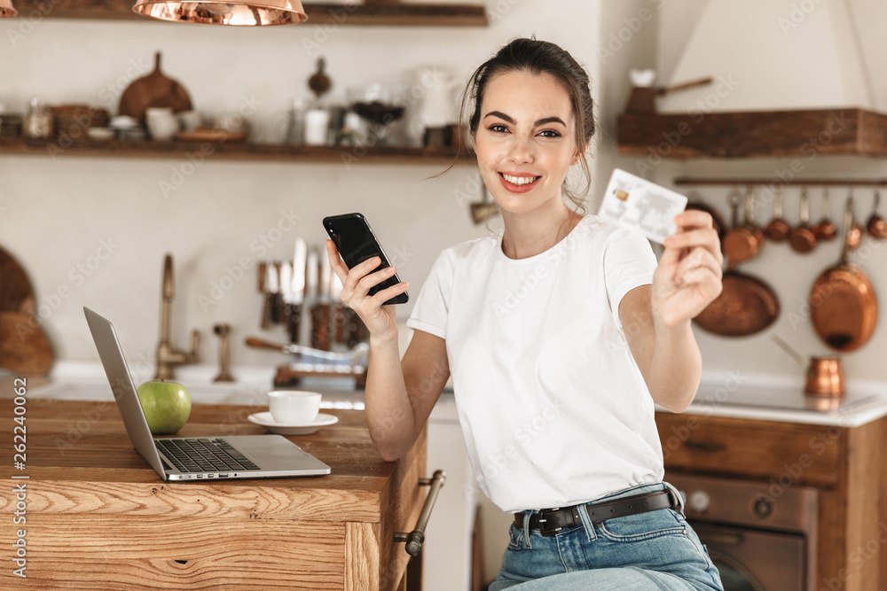 Young student girl sitting indoors using laptop computer chatting by mobile phone holding credit card. Young student girl sitting indoors using laptop computer chatting by mobile phone holding credit card.