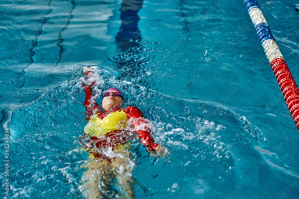 Female athlete in a red-yellow swimsuit is swimming on his back. Splashes of water scatter in different directions.