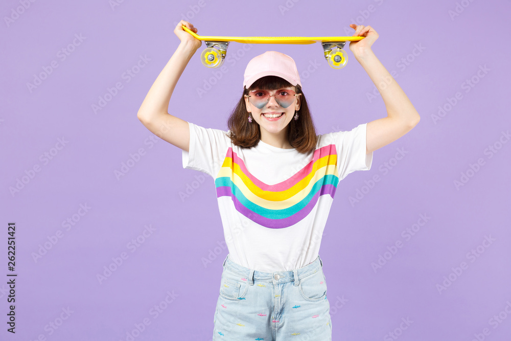 Smiling pretty teen girl in vivid clothes heart eyeglasses holding yellow skateboard over head isolated on violet pastel wall background. People sincere emotions lifestyle concept. Mock up copy space. Smiling pretty teen girl in vivid clothes heart eyeglasses holding yellow skateboard over head isolated on violet pastel wall background. People sincere emotions lifestyle concept. Mock up copy space.
