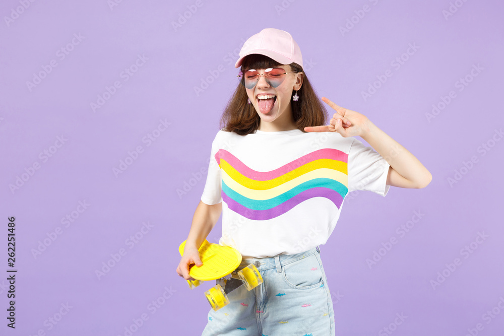 Crazy teen girl with skateboard depicting heavy metal rock sign, showing tongue keeping eyes closed isolated on violet pastel background. People sincere emotions lifestyle concept. Mock up copy space. Crazy teen girl with skateboard depicting heavy metal rock sign, showing tongue keeping eyes closed isolated on violet pastel background. People sincere emotions lifestyle concept. Mock up copy space.