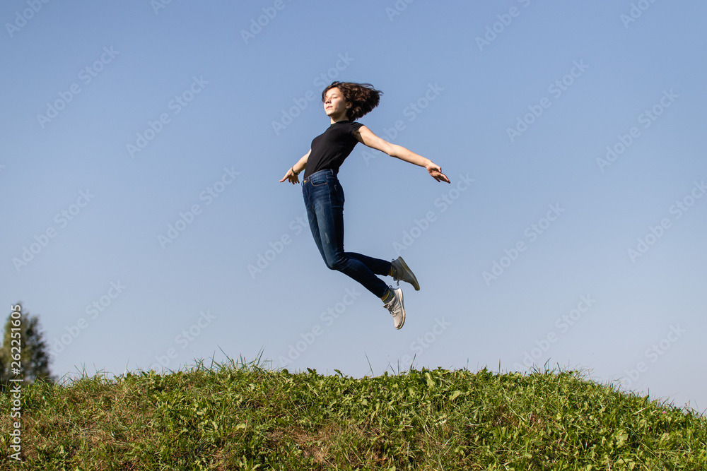 slim teen girl dressed in jeans and a black top jumping high over green grass against the sky. moment of flight slim teen girl dressed in jeans and a black top jumping high over green grass against the sky. moment of flight