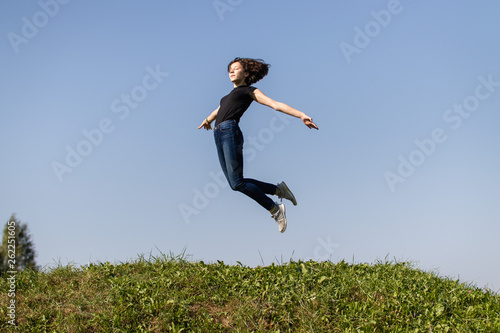 slim teen girl dressed in jeans and a black top jumping high over green grass against the sky. moment of flight