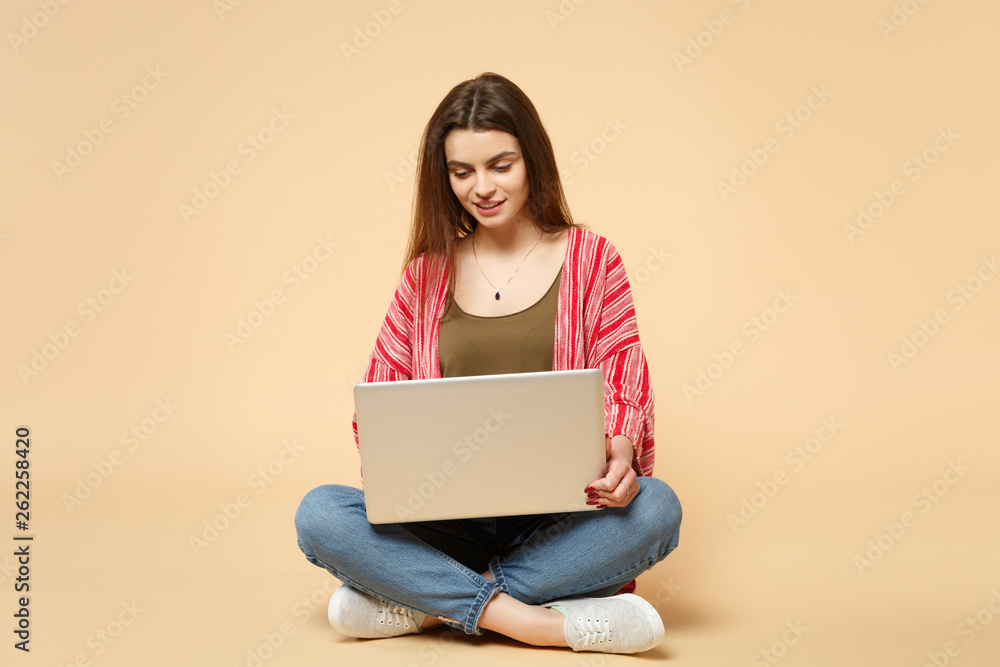 Naklejka premium Portrait of beautiful young woman in casual clothes sitting, using laptop pc computer isolated on pastel beige wall background in studio. People sincere emotions lifestyle concept. Mock up copy space.