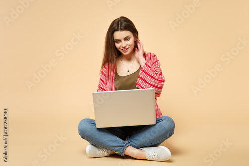 Portrait of excited young woman in casual clothes sitting, using laptop pc computer isolated on pastel beige wall background in studio. People sincere emotions lifestyle concept. Mock up copy space.
