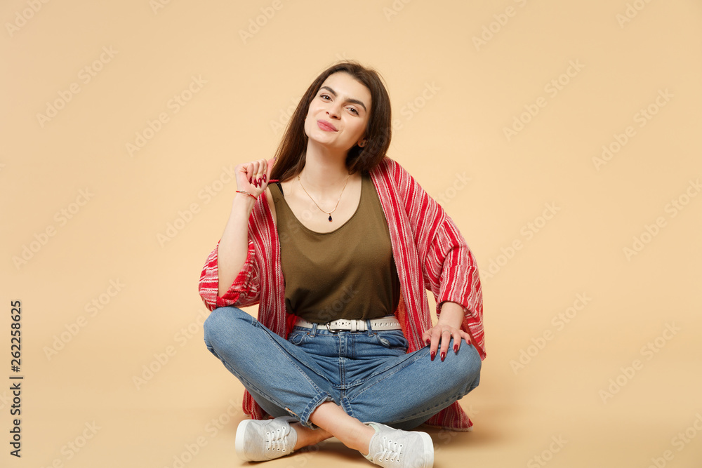 Portrait of smiling attractive young woman in casual clothes sitting, looking camera isolated on pastel beige wall background in studio. People sincere emotions, lifestyle concept. Mock up copy space.
