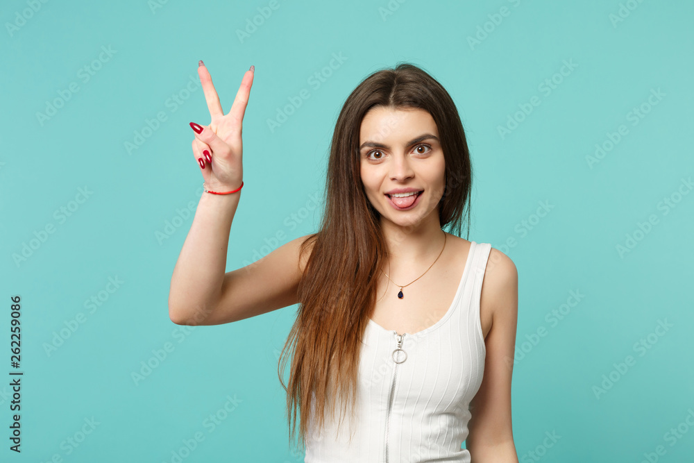 Portrait of beautiful young woman in light casual clothes showing tongue, victory sign isolated on blue turquoise background in studio. People sincere emotions, lifestyle concept. Mock up copy space. Portrait of beautiful young woman in light casual clothes showing tongue, victory sign isolated on blue turquoise background in studio. People sincere emotions, lifestyle concept. Mock up copy space.