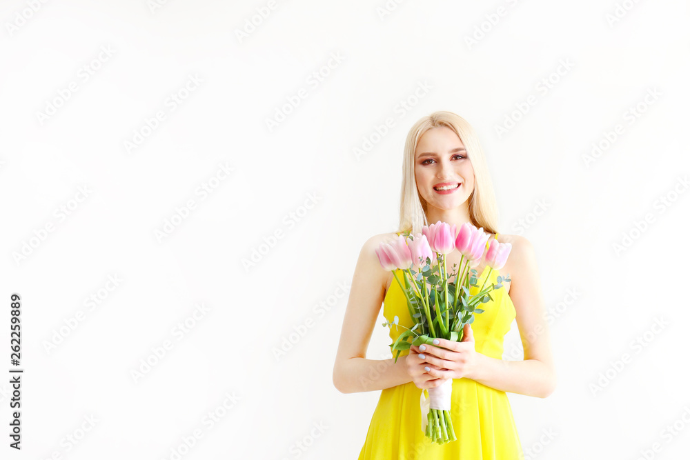 Studio portrait of gorgeous young blonde woman with long straight hair wearing bright yellow dress and holding bouquet of many tender pink tulips. Gray isolated background, copy space, close up. Studio portrait of gorgeous young blonde woman with long straight hair wearing bright yellow dress and holding bouquet of many tender pink tulips. Gray isolated background, copy space, close up.