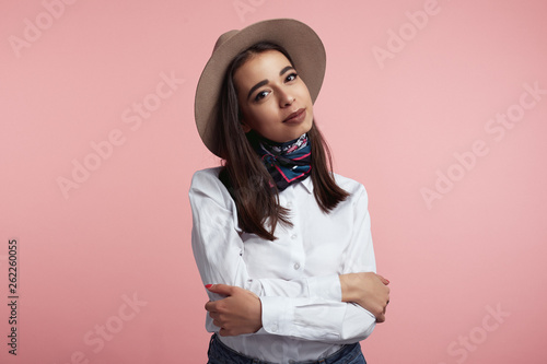 Confident pretty young girl with crossed hands, dressed in elegant hat, white shirt and stylish scarf, isolated over pink backdrop