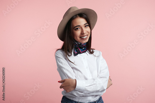 Young beautiful brunette girl wearing elegant hat, white shirt and stylish scarf, standing with hands crossed against pink wall