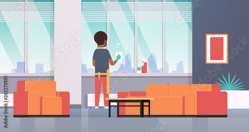 man in gloves and apron cleaning windows with rag cleaner spray rear view african american guy doing housework concept modern apartment living room interior flat full length horizontal