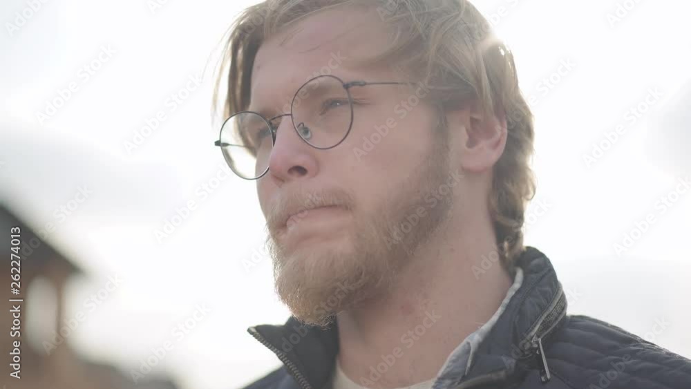 Portrait of the blond man drinking beer outdoors looking away. Bearded man in glasses enjoying his alcohol drink, hair fluttering in the wind. Portrait of the blond man drinking beer outdoors looking away. Bearded man in glasses enjoying his alcohol drink, hair fluttering in the wind.