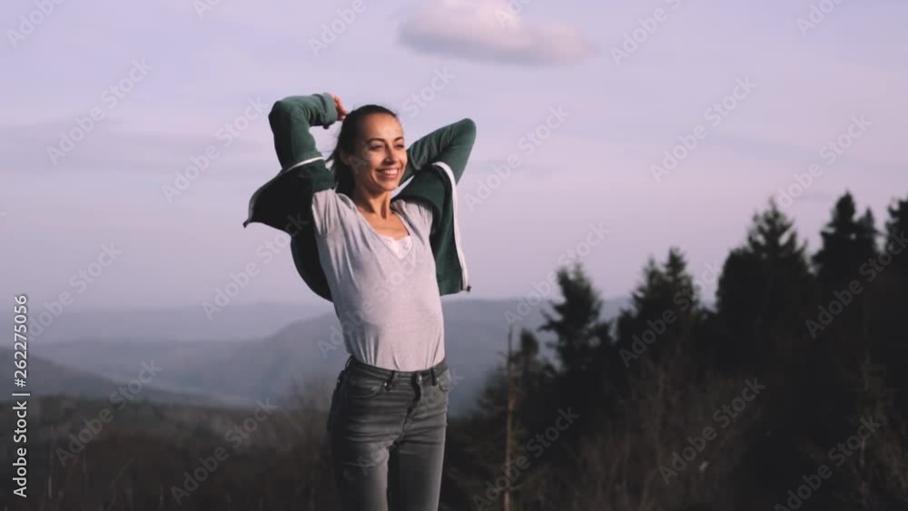 young female hiker is standing raising hands on edge of mountain ridge against background of sunset sky. Woman is hiking in mountains before sunset. strong wind is fluttering woman's hair and shirt