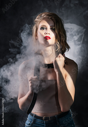 Conceptual photo of a young girl with disheveled wet hair, naked shoulders, aggressive make-up, and a men's leather belt around her neck on a gray background in artistic shadows and smoke
