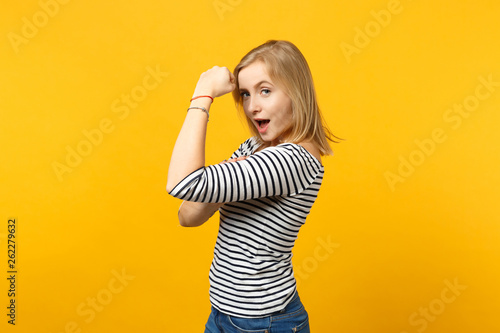 Side view of funny young woman in striped clothes looking camera showing biceps, muscles isolated on yellow orange background in studio. People sincere emotions, lifestyle concept. Mock up copy space.