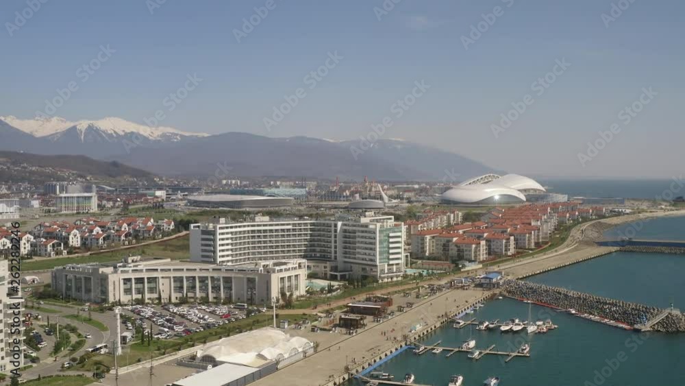 Modern olympic city on sea shore and snowy mountain background. Aerial footage modern architecture in developed city on blue sea shore and mountain landscape on skyline. Modern olympic city on sea shore and snowy mountain background. Aerial footage modern architecture in developed city on blue sea shore and mountain landscape on skyline.