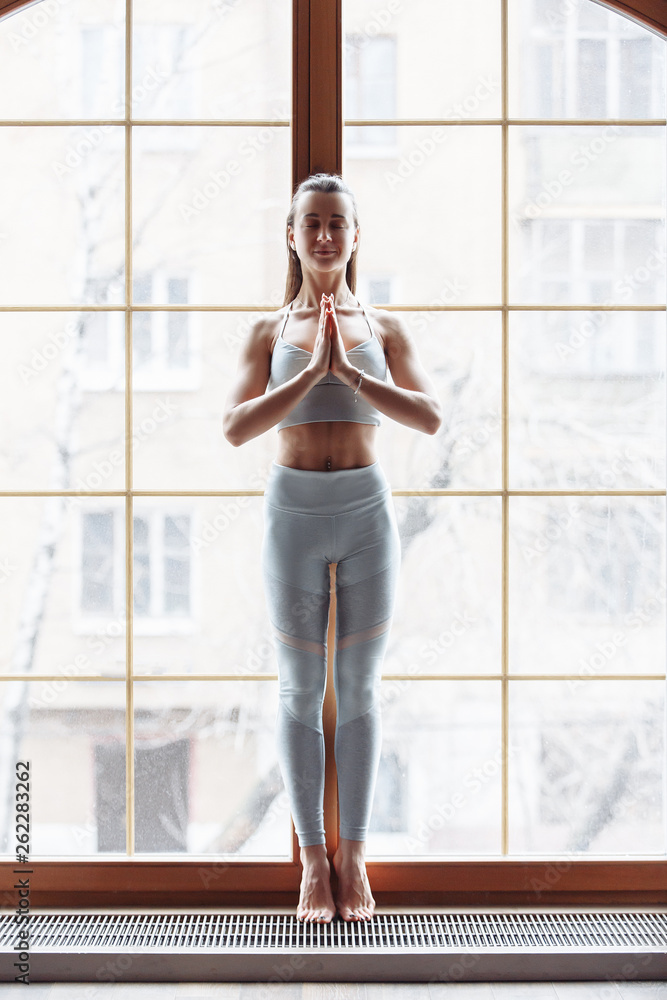 Fototapeta premium Slim beautiful young girl in sportswear is standing at the window and meditating with closed eyes. Concept of relaxation and spirituality. Yoga concept