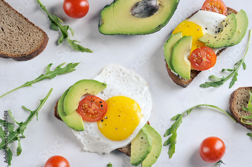 Toasts with avocado, cherry tomato and fried egg. The composition is supplemented with arugula greens. Light background. View from above.