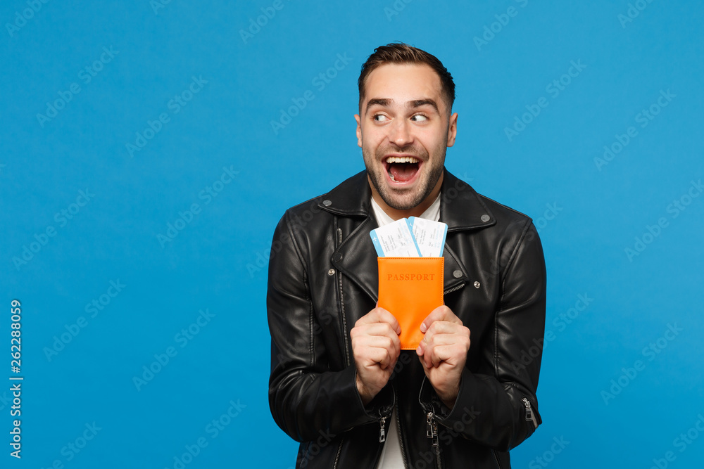 Young happy unshaven man in black jacket white t-shirt hold in hand passport tickets isolated on blue wall background. Passenger traveling abroad getaway. Air flight journey concept Mock up copy space Young happy unshaven man in black jacket white t-shirt hold in hand passport tickets isolated on blue wall background. Passenger traveling abroad getaway. Air flight journey concept Mock up copy space