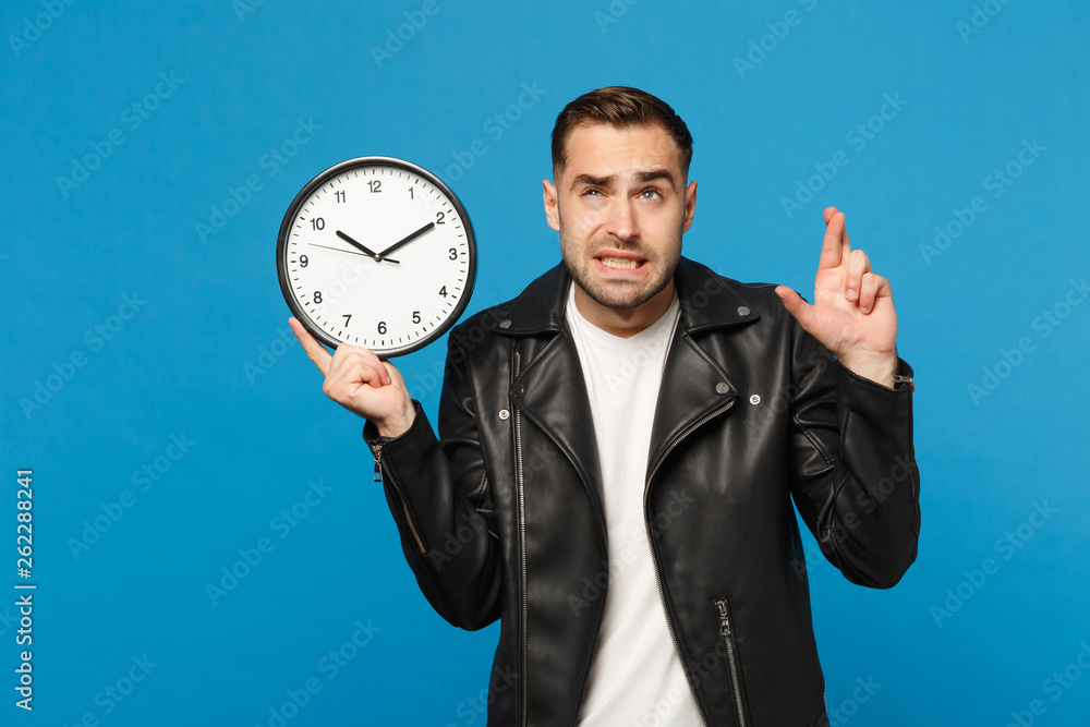 Sad upset stylish young unshaven man in black leather jacket white t-shirt holding round clock isolated on blue wall background studio portrait. People lifestyle concept. Hurry up. Mock up copy space.