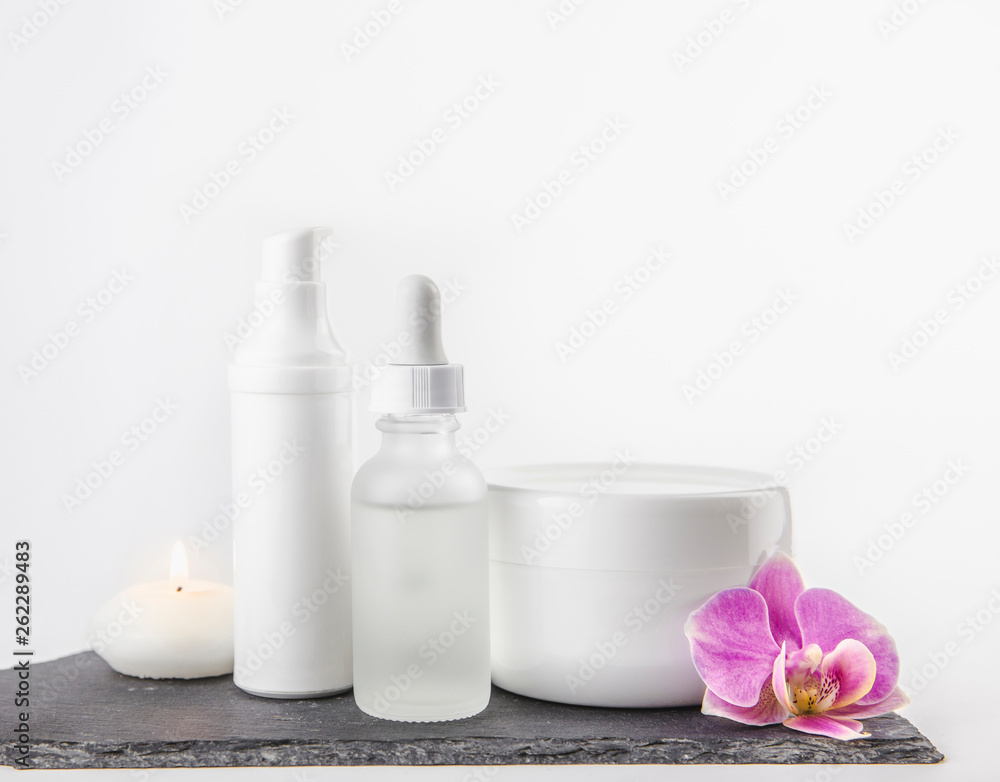 Different material shape and size cosmetic product cream, oil containers on white background standing on black stone base, pink orchid blossom for decoration. Lot of copy space. Different material shape and size cosmetic product cream, oil containers on white background standing on black stone base, pink orchid blossom for decoration. Lot of copy space.