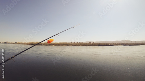 Fishing rod with a bright orange large float. Background river, ships, houses and mountains on the opposite shore. Bright sun. 