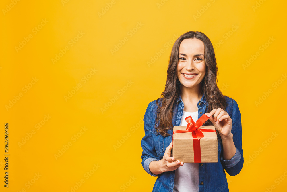 Smiling young beautiful woman holds red gift box. Isolated over yellow background. Studio. Smiling young beautiful woman holds red gift box. Isolated over yellow background. Studio.