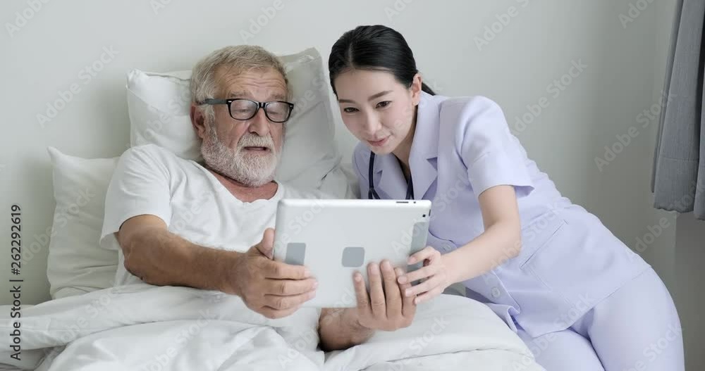 Nurse with senior patient in bedroom. Asian female nurse with bearded white male in bed. Using tablet together. Senior lifestyle concept. Nurse with senior patient in bedroom. Asian female nurse with bearded white male in bed. Using tablet together. Senior lifestyle concept.