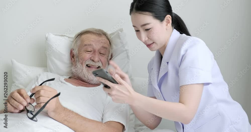 Nurse with senior patient in bedroom. Asian female nurse with bearded white male in bed. Looking at their photos. Senior lifestyle concept. Nurse with senior patient in bedroom. Asian female nurse with bearded white male in bed. Looking at their photos. Senior lifestyle concept.
