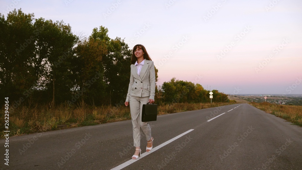 business woman with black briefcase walks in light suit and white high-heeled shoes goes outside city along asphalt with white markings, view from front