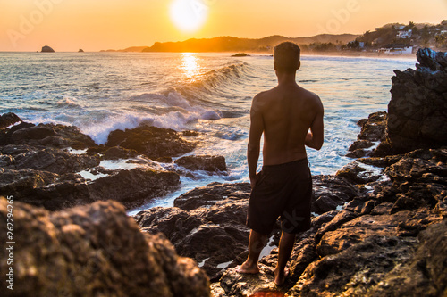 Homme torse nu regardant les vagues en couché de soleil debout sur un rocher en face de la baie de Zipolite. 