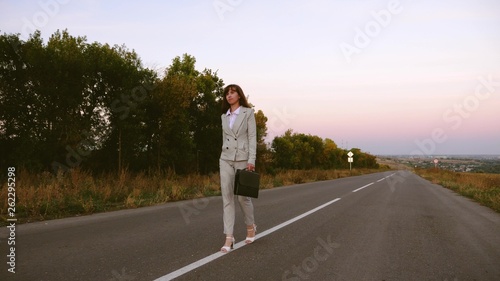business woman with black briefcase walks in light suit and white high-heeled shoes goes outside city along asphalt with white markings  view from front