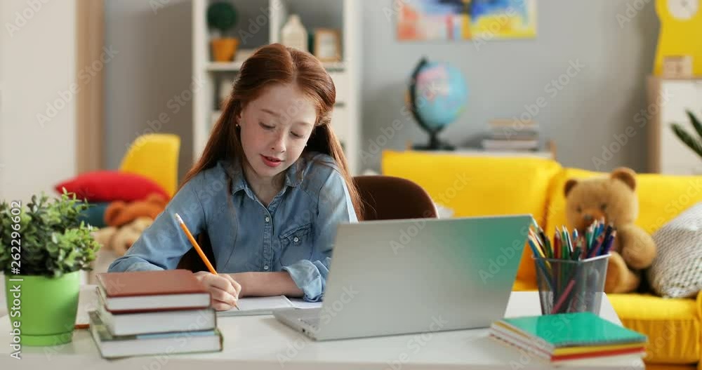 Caucasian schoolgirl with red long hair writing something while doing homework at the table with laptop computer in the cozy room. Caucasian schoolgirl with red long hair writing something while doing homework at the table with laptop computer in the cozy room.