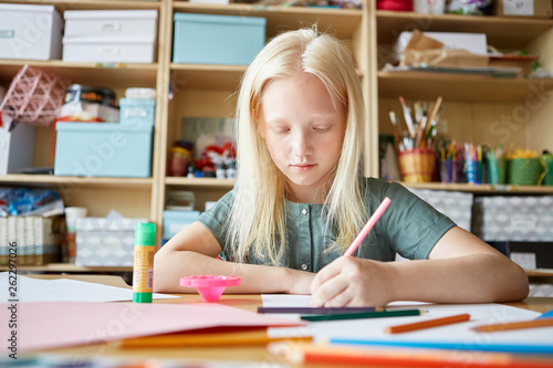 Lovely albino girl drawing good picture with colored pencils while sitting at table during art lesson at school
