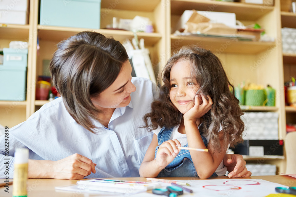 Fototapeta premium Młody nauczyciel uśmiecha się i patrzy na wesołą dziewczynę podczas wspólnego malowania podczas lekcji w szkole artystycznej