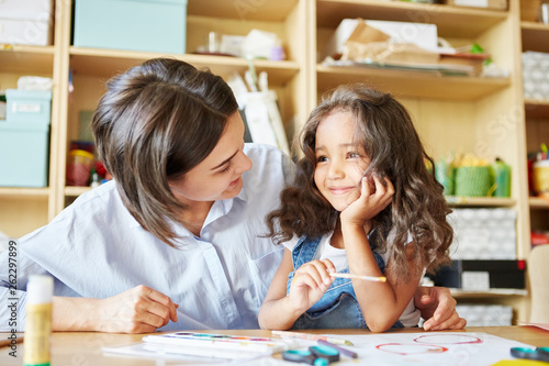 Young teacher smiling and looking at cheerful girl while painting together during lesson in art school