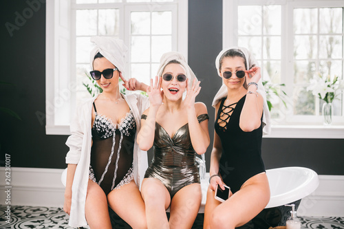 Theme female friendship. Party girlfriends, relaxation and spa procedures, hen party. Three Caucasian beautiful women sitting inside on bathroom in sunglasses, head towel and bathrobe, underwear