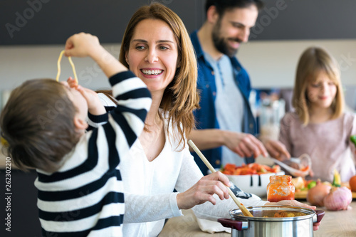 Cute little boy having fun with his young mother while eating spaghetti on kitchen at home.