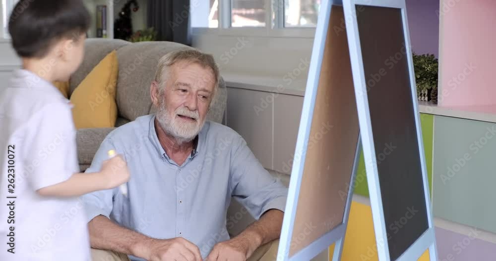 Senior man with his grandson using white board. White bearded man with asian boy in living room. Teaching grandpa what he is drawing. Senior man with his grandson using white board. White bearded man with asian boy in living room. Teaching grandpa what he is drawing.