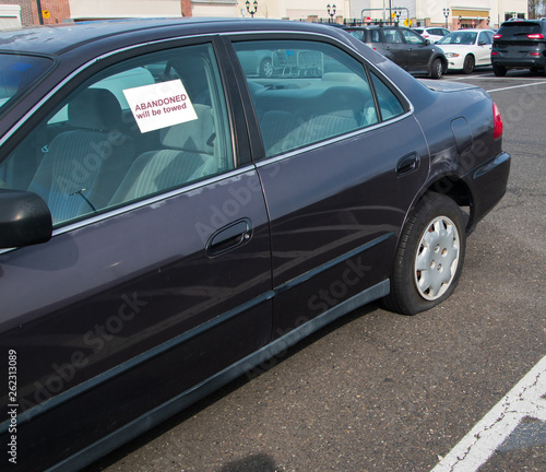 Old car with flat tire in parking lot with sticker on window that says Abandoned.