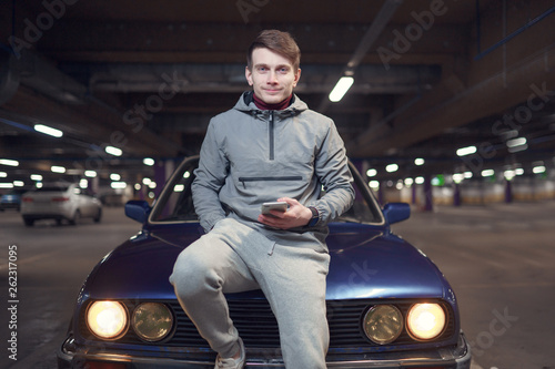 Image of man with phone sitting on hood of black car. © Sergey