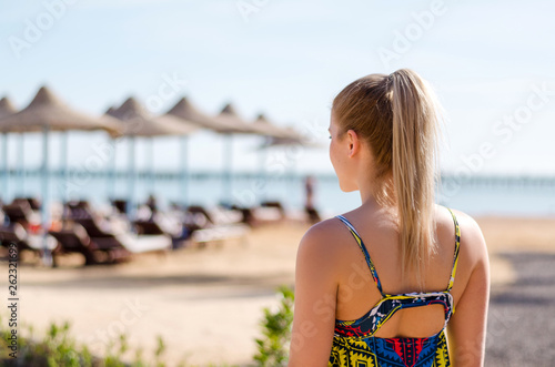 Young woman with ponytail, blonde hair is looking on the sea horizon, vacation mood, summer travel concept. Beach umbrellas on the background