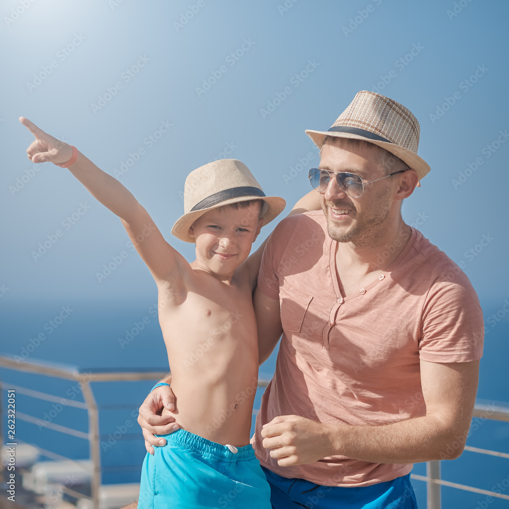 European son and father  in sun hats enjoying their summer vacation against blue sea.